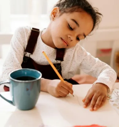 A child drawing at a kitchen table with a mug of drink next to her.