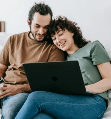 A young couple sat down looking at a laptop together and smiling.